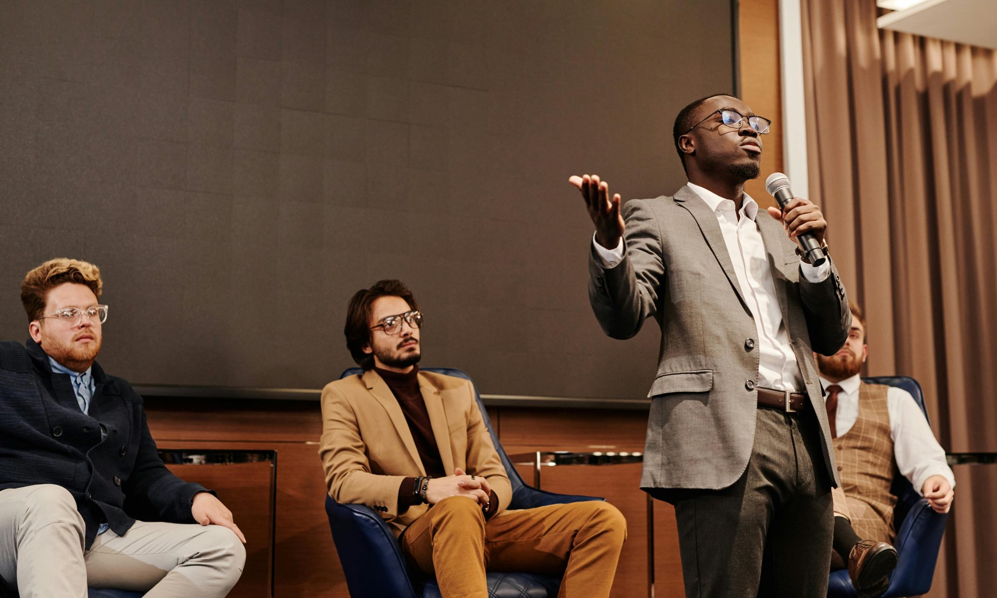 Businessman stands with a microphone while colleagues sit behind him