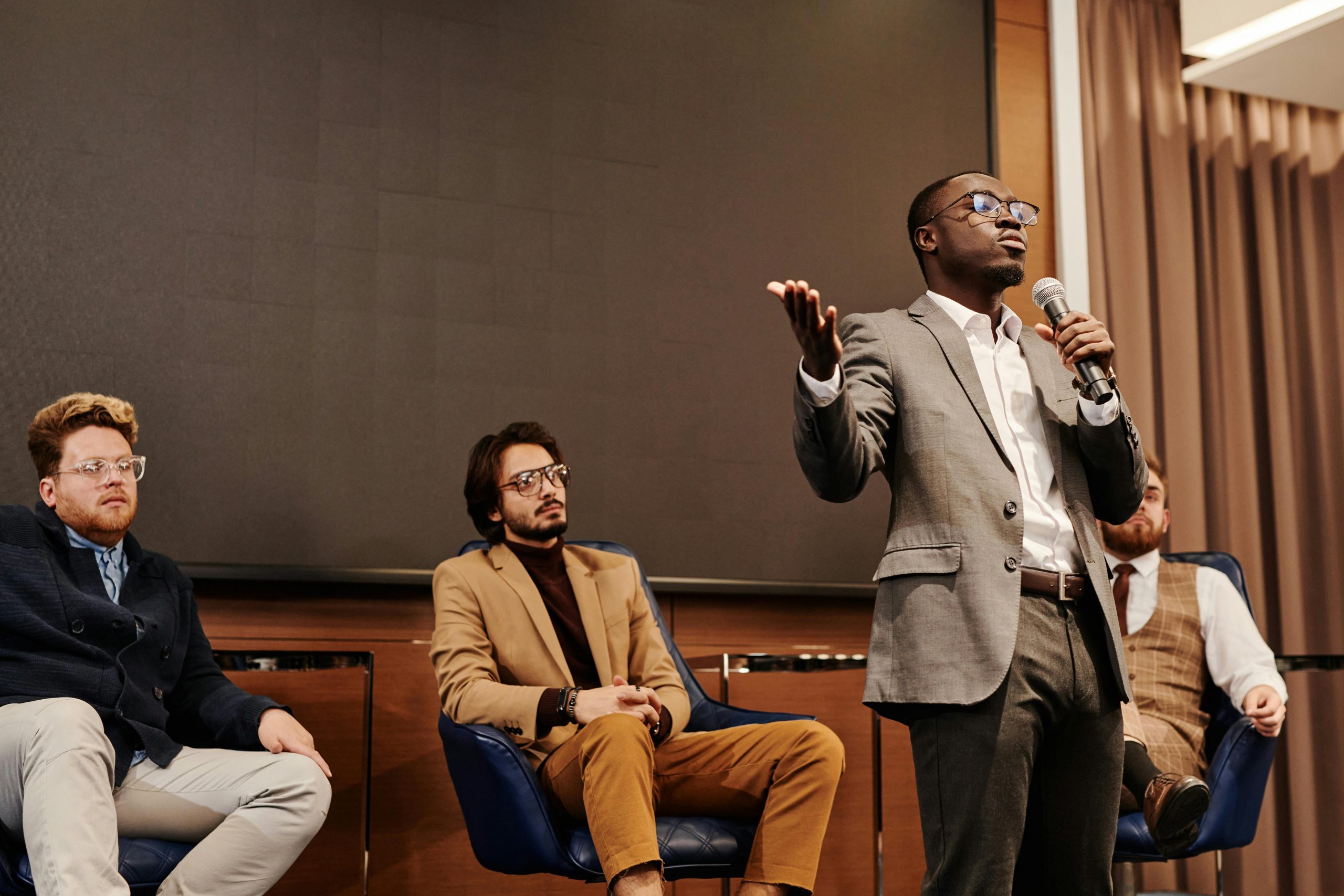 Businessman stands with a microphone while colleagues sit behind him